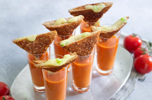 Small glasses of tomato soup topped with a grilled brie sandwich wedge